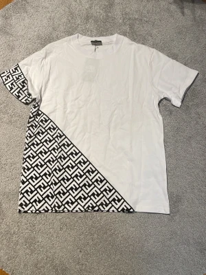 Vit t-shirt med svart grafiskt mönster - Säljer en vit t-shirt från Fendi med korta ärmar och ett fett svart grafiskt mönster på ena ärmen och nedre delen av tröjan. Materialet är mjuk bomull och passformen är normal. Perfekt för dig som gillar att sticka ut med designerplagg.