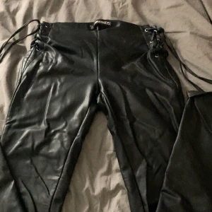 Svarta bootcut skinnbyxor med snörning - Säljer ett par svarta byxor i skinnimitation med snörning i sidorna och vida ben. Byxorna har en cool look med släta ytor och långa snören som detalj. Perfekta för dig som vill sticka ut och gillar edgy stil. 