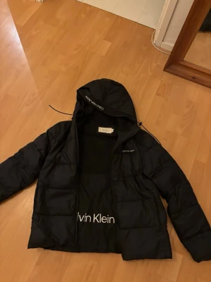 Svart pufferjacka Calvin Klein Jeans - Svart pufferjacka från Calvin Klein Jeans med huva och dragkedja. Jackan har broderad logga på bröstet och huvan, samt quiltad design. Perfekt för kalla dagar och har en clean, modern look som funkar till streetwear.