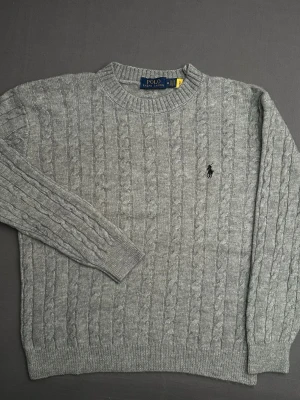Grå kabelstickad tröja Polo Ralph Lauren - Snygg grå kabelstickad tröja från Polo Ralph Lauren i storlek M. Tröjan har rund hals, långärmade ribbade muddar och den klassiska broderade loggan på bröstet. Perfekt för dig som gillar stilrena och tidlösa plagg med premiumkänsla.