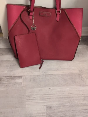 Röd axelväska från DKNY med dragkedja - Stilren röd axelväska från DKNY i skinn med guldiga detaljer och dragkedjor på sidorna. Väskan har två handtag, medföljande matchande clutch och rymligt brunt innerfoder. Perfekt för dig som vill ha en snygg och praktisk väska med exklusiv känsla.