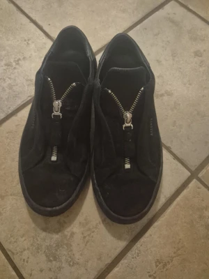 Svarta sneakers från Axel Arigato EU39 - Säljer ett par svarta sneakers från Axel Arigato i mocka med cool dragkedjedetalj framtill. Skorna har platt sula och rund tå, och är handgjorda i Portugal. Stilren design med diskret logga på sidan. Passar dig som gillar minimalistisk look och snygga detaljer.