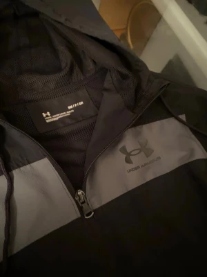  Under Armour Vindjacka  - Svart vindjacka från Under Armour med grå panel över bröstet och logga. Jackan har huva med snörning, hel dragkedja och meshfoder på insidan. Perfekt för dig som gillar sportig stil och vill ha något lätt att slänga på sig.