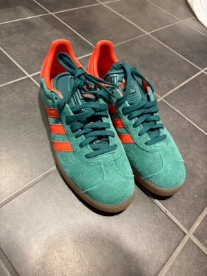Adidas Gazelle turkos/röda sneakers - Säljer ett par Adidas Samba sneakers i turkos mocka med rödaoranga detaljer och klassiska tre ränder på sidan. Skorna har snörning, gummisula och rund tå. Perfekt för dig som vill sticka ut med färg! Aldrig använda! 