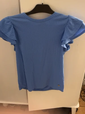 Blå ribbad topp från Pernilla Wahlgren - Säljer en blå ribbad topp från Pernilla Wahlgren i storlek XS. Toppen har korta volangärmar och är tillverkad i ett mjukt material som känns skönt mot huden. Perfekt för dig som gillar en enkel men ändå söt stil med lite extra detaljer på ärmarna. Använd 1 gång