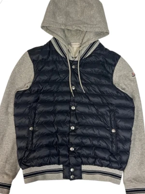 Moncler cardigan med huva - Snygg cardigan från Moncler med mörkblå quiltad front och gråa ärmar samt huva i sweatshirtmaterial. Jackan har knappar framtill, ribbade muddar och snörning vid huvan. Perfekt för dig som vill ha en sportig och trendig look. Storlek M men passar xs och den är självklart äkta. Hör av dig vid frågor. Kan sänka vid snabb affär