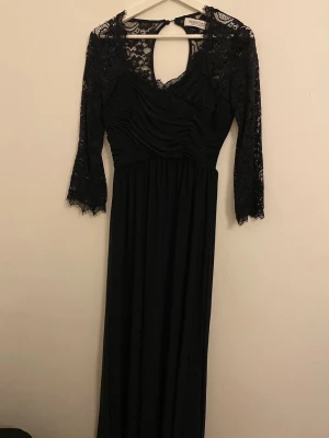 Svart långklänning med spets från Chiara Forti - Svart långklänning med spetsdetaljer på ärmar och axlar. Klänningen har v-ringning, rynkad överdel och trekvartsärmar i spets. Perfekt för fester och ger en elegant look med öppen rygg och utsvängande nederdel.