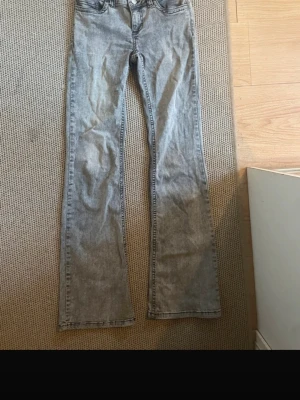 Grå bootcut jeans från Nelly - Säljer ett par grå bootcut jeans från Nelly med snygg tvättad look och svarta broderade detaljer på bakfickorna. Jeansen har hög midja, dubbla knappar och klassisk femficksdesign. Materialet är jeans med en mjuk känsla och passformen är loose.