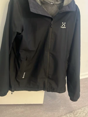 Svart vindjacka från Haglöfs Gore-Tex - Svart vindjacka från Haglöfs med Gore-Tex-material som håller dig torr. Jackan har dragkedja framtill, två sidofickor med dragkedja och hög krage. Perfekt för dig som gillar att vara ute oavsett väder. Snygg och funktionell design med diskret logga på bröstet.