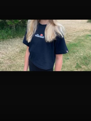 Svart Ellesse t-shirt med broderad logga - Svart t-shirt från Ellesse i storlek EU 34. Klassisk modell med rund hals och korta ärmar. Framsidan har en broderad Ellesse-logga i vitt, orange och rött. Materialet är mjuk bomull som känns skön mot huden. Perfekt till jeans eller shorts för en chill look.