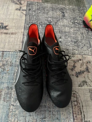 Svarta Puma fotbollsskor med orange detaljer - Säljer ett par svarta fotbollsskor från Puma med orange detaljer och innersida. Skorna har snörning, mönstrad sula med dobbar och en vit Puma-logga på sidan. Ovandelen har hexagonmönster och materialet är syntet. Perfekta för dig som vill sticka ut på planen. Använda 1 gång