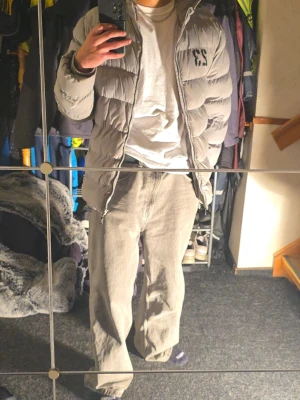 Grå pufferjacka från Vinson - Grå pufferjacka från Vinson med huva och dragkedja. Jackan har tryck med siffran 23 och Vinson Original-logga på bröstet, med defekter. Fylld med syntetmaterial och har långa ärmar. Perfekt för kalla dagar och har en clean, sportig vibe. Slitage finns även på vänster arm.