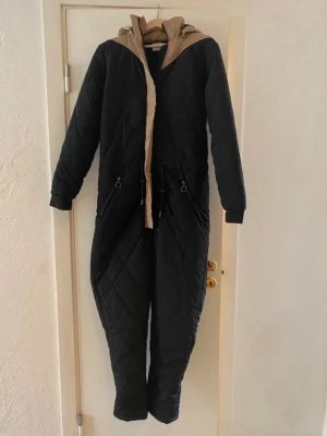 Quiltad svart overall från KA:NT - Quiltad svart overall från KA:NT Copenhagen med beige detaljer och huva. Overallen har dragkedja framtill, snörning i midjan och två fickor med dragkedja. Perfekt för kalla dagar när du vill vara både varm och stilren.