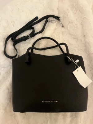 Svart handväska från Charles & Keith - Stilren svart handväska från Charles & Keith i strukturerat syntetmaterial. Väskan har både handtag och en avtagbar axelrem, samt dragkedja upptill. Insidan är ljus med flera fack för smart organisering. Perfekt storlek för allt du behöver ha med dig. (Aldrig använd)