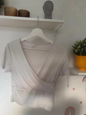 Ljus ribbad omlottopp - Säljer en ljus, ribbad omlottopp med korta ärmar och v-ringad hals. Toppen har ett stilrent omlott-snitt och är tillverkad i mjukt material som känns skönt mot huden. Perfekt för dig som gillar enkla och snygga basplagg med lite extra detalj.