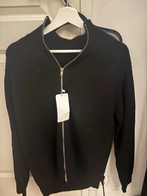 Svart stickad kofta från Maison Margiela - Svart stickad kofta från Maison Margiela med ribbad struktur och hel dragkedja framtill. Modellen har hög krage och långa ärmar, perfekt för dig som gillar minimalistisk och stilren design. Snygg att bära öppen eller stängd beroende på humör.