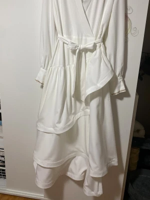 Vit omlottklänning med volanger - Säljer en elegant vit långklänning med omlott-design och knytband i midjan. Klänningen har långa ärmar med diskreta detaljer vid manschetterna och flera lager volanger i kjolen som ger en snygg och flödande siluett. Perfekt för dig som gillar stilrena och romantiska plagg. Använd en gång till studenten bara. 