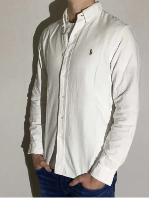Vit skjorta från Polo Ralph Lauren - Vit skjorta från Polo Ralph Lauren i slim fit med klassisk krage och knappar framtill. Skjortan har lång ärm och den ikoniska broderade loggan på bröstet. Tillverkad i mjuk bomull som känns skön mot huden. Perfekt för dig som gillar stilrena och tidlösa plagg.