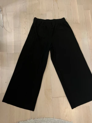 Svarta vida plisserade byxor - Säljer ett par svarta byxor med plisserad struktur och breda ben. De har elastisk midja och är riktigt sköna att ha på sig. De är även fladdriga och luftiga och därför perfekta till sommaren och våren. Passar dig som gillar loose fit och vill ha en chill och trendig look. Materialet känns mjukt och lätt. Storlek 146 och passar 10-12 åringar.