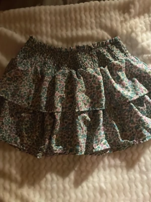 Blommig volangkjol med resår - Säljer en söt kort kjol med blommigt mönster i blått, rosa och grönt. Kjolen har bred resår i midjan och flera lager volanger som ger en cool och lekfull look. Perfekt för dig som gillar färg och mönster!