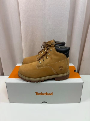 Timberland boots i mocka - Klassiska Timberland boots i beige mocka med bruna snören, guldfärgade öljetter och vadderad svart krage. Grov sula för extra grepp och slitstyrka. Perfekta för höst och vinter, med snygg kontrastsöm och ikonisk Timberland-logga på sidan.