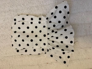 Vit prickig bandeau topp - Säljer en vit bandeau topp med svarta prickar och volang nedtill. Toppen är axelbandslös och har en smockad baksida för extra stretch. Perfekt för dig som gillar en lekfull och trendig stil. Materialet känns som bomull och toppen har en croppad passform.