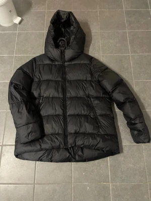 Svart pufferjacka med huva - Säljer en svart pufferjacka med stor huva och dragkedja framtill. Jackan har två snedställda fickor med dragkedja och en quiltad design som ger en riktigt schysst look. Perfekt för kalla dagar när du vill hålla dig varm och stilsäker.