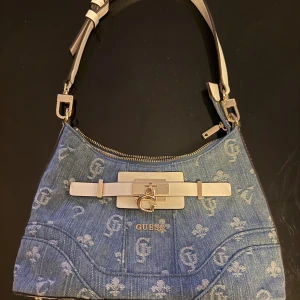 Blå axelväska i denim från Guess - Snygg axelväska från Guess i blå denim med ljusa logomönster över hela väskan. Väskan har vita detaljer i syntet, guldfärgade metalldetaljer och dragkedja upptill. Insidan är fodrad med beige tyg och har innerfack med dragkedja. Perfekt för att lyfta din outfit.