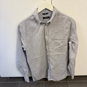 Ljusgrå skjorta från J.Lindeberg - Säljer en stilren ljusgrå skjorta från J.Lindeberg i storlek S. Skjortan har klassisk krage, knappstängning framtill och en bröstficka. Tillverkad i mjuk bomull och passar perfekt till jeans eller chinos.