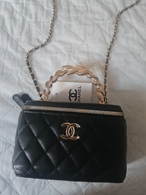 Svart quiltad handväska - Säljer en svart quiltad handväskal med guldfärgad kedja och flätat handtag. Väskan har dragkedja, ikonisk CC-logga framtill och röd insida med spegel i locket. Perfekt storlek för det viktigaste och riktigt snygg detalj till din outfit.
