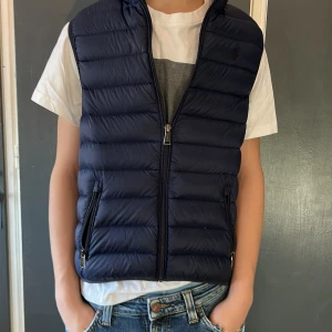Mörkblå dunväst från Polo Ralph Lauren - Snygg mörkblå dunväst från Polo Ralph Lauren med quiltad design och hög krage. Västen har två fickor med dragkedja och stängs med en dragkedja framtill. Perfekt att slänga över en hoodie eller t-shirt för en chill look. Det är m i barn storlek asså 10-12 jag ör 160 å ni ser hur den sitter på mig