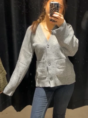 Grå kofta med knappar - Mysig grå kofta med v-ringning och stora knappar framtill. Koftan har två praktiska fickor och är stickad i ett mjukt material. Perfekt för lager-på-lager och passar till jeans eller kjol.