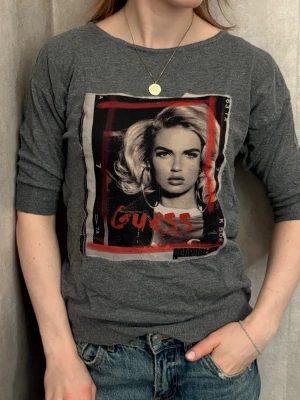 Grå t-shirt med tryck från Guess - Grå t-shirt från Guess med coolt fototryck framtill i svartvitt och röda detaljer. Modellen har halvlånga ärmar och rund halsringning. Materialet är mjukt och stretchigt, perfekt till jeans eller kjol.