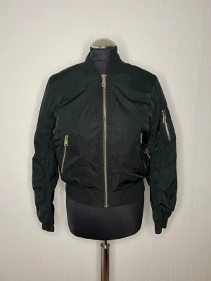 Svart bomberjacka från Topshop Petite - Säljer en svart bomberjacka från Topshop Petite i storlek 36. Jackan har klassisk bombermodell med ribbade muddar, två fickor med dragkedja framtill och en extra ficka på ärmen. Tillverkad i polyester och stängs med en grov dragkedja i silver.