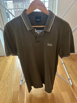 Olivgrön piké från Hugo Boss L - Snygg olivgrön pikétröja från Hugo Boss med vit logga på bröstet och kontrasterande vita detaljer på krage och ärmslut. Klassisk pikékrage med knappar framtill. Perfekt till jeans eller shorts för en clean look.