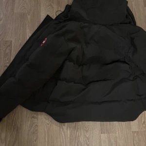 Mörkgrå pufferjacka från Canada Goose - Säljer en mörkgrå pufferjacka från Canada Goose med huva och klassisk logga på ärmen. Jackan har dragkedja, stora fickor och ribbade muddar vid ärmsluten. Perfekt för kalla dagar och riktigt snygg till streetwear-stilen. Vid snabb affär kan vi diskutera pris!