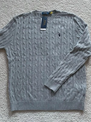 Grå kabelstickad tröja Polo Ralph Lauren - Säljer en grå kabelstickad tröja från Polo Ralph Lauren i 100% bomull. Klassisk rund halsringning, ribbade muddar och ikoniska broderade loggan på bröstet. Perfekt till lager på lager under höst och vinter.