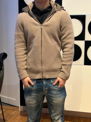 Massimo Dutti cardigan  - Säljer en beige stickad jacka från Massimo Dutti med ribbad struktur och dragkedja framtill. Jackan har en vadderad huva i matchande färg och långa ärmar. Perfekt för dig som gillar en clean och chill stil.