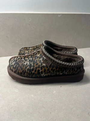 UGG leopardmönstrade slip-ins - Säljer ett par UGG slip-ins med leopardmönster i bruna och beige toner. Skorna har en mjuk, fluffig insida och grov sula. Dekorativt band runt öppningen och rund tå. Perfekta för dig som vill ha något både bekvämt och snyggt hemma.