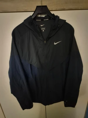 Nike miller jacka - Säljer en snygg tvåfärgad vindjacka från Nike i mörkblått och grått. Jackan har huva med dragsko, hel dragkedja framtill och reflekterande detaljer. Perfekt för blåsiga dagar och har meshfoder på insidan för extra komfort.