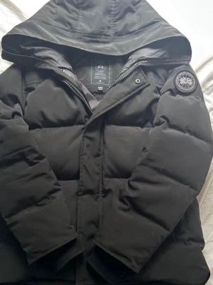 Svart pufferjacka från Canada Goose - Säljer en svart pufferjacka från Canada Goose med huva och patch på ärmen. Jackan har dragkedja och knappar framtill, samt två sidofickor. Perfekt för kalla dagar och riktigt skön att bära. Klassisk och stilren modell som funkar till allt.