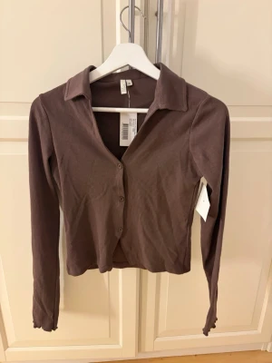 Brun ribbad kofta från NLY Trend - Brun ribbad kofta från NLY Trend i storlek M. Tröjan har långa ärmar, klassisk krage och knäppning framtill med fyra knappar. Materialet är mjukt och stretchigt, perfekt för lager på lager under höst och vår.