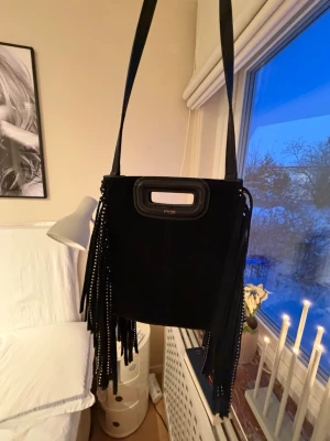 Svart axelväska med fransar från Maje - Säljer en svart axelväska från Maje i mocka med långa fransar på sidorna och coola nitar. Väskan har både axelrem och ett handtag i toppen. Den är super fin och knappt använt! Pris kan diskuteras😊💕