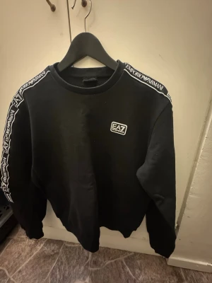 Svart EA7 Emporio Armani sweatshirt - Svart sweatshirt från EA7 Emporio Armani med logga på bröstet och tydliga logoband längs axlarna. Tröjan har rund halsringning och långa ärmar. Perfekt för dig som gillar stilrena och sportiga plagg med premiumkänsla.