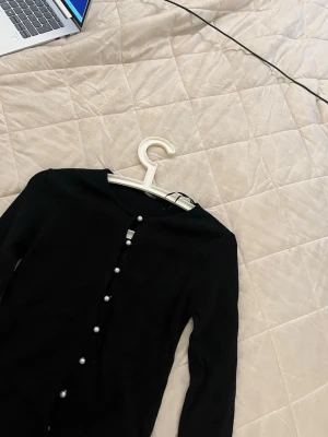 Svart ribbad kofta med pärlknappar - Svart jättefin kofta med pärlknappar från Gina Tricot! 🤍I strl L men passar även M. i nytt skick då jag endast använt en gång!          Nypris: 500kr