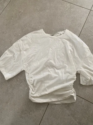 Vit t-shirt med snörning från Gina Tricot - Säljer en vit t-shirt från Gina Tricot i storlek S. T-shirten har korta ärmar och en snygg snörningsdetalj i sidan som ger en cool twist. Tillverkad i mjuk bomull och passar perfekt till jeans eller kjol.