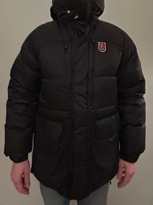 Svart dunjacka från Fjällräven, stl S (man) - Säljer en äkta, svart, lång och riktigt varm dunjacka från Fjällräven.  Jackan har huva med snörning, stora fickor framtill, gul dragkedja och Fjällräven-logga på bröstet. Jackan har två små lagningar ( ser bilder ) storlek S (man) kan gå som unisex och är då ca stl L/40 i dam. Rök- och djufritt hem.