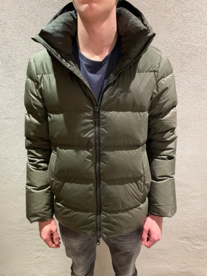 Olivgrön pufferjacka från J.Lindeberg - Säljer en olivgrön pufferjacka från J.Lindeberg med hög krage och huva. Jackan har en clean look med diskret logga på bröstet, dragkedja framtill och två sidofickor. Perfekt för kalla dagar och riktigt skön att bära.