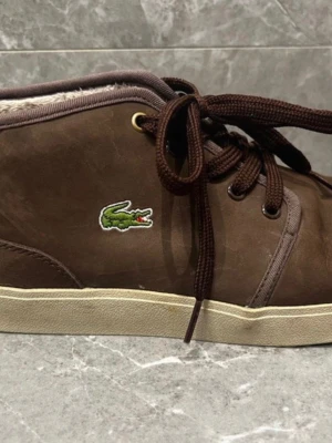 Bruna mockasneakers från Lacoste - Bruna sneakers från Lacoste i mjuk mocka med vitt foder och klassisk krokodillogo på sidan. Skorna har snörning, beige sula och rund tå. Perfekta för höst och vinter med sitt varma, fluffiga innerfoder.