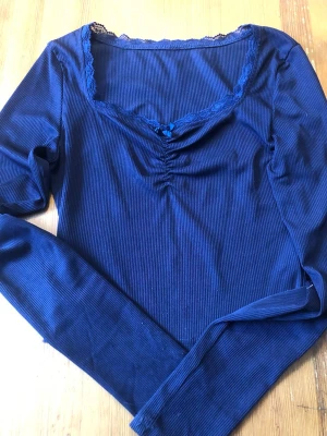 Marinblå ribbad topp med spets - Säljer en marinblå långärmad topp med ribbad struktur. Toppen har spetsdetaljer längs ringningen, en liten rosett. Väldigt fint skick skicka om vid intresse 